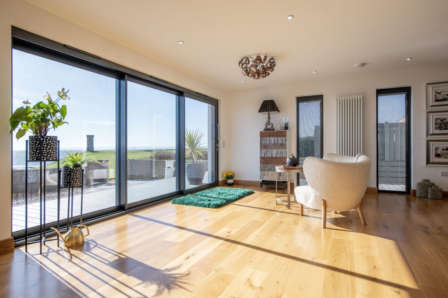 Bright, modern living room with large glass doors and bespoke glass solutions overlooking a grassy landscape and sea. Wooden floors, indoor plants, a cozy chair, and a small green rug decorate the sunlit space.