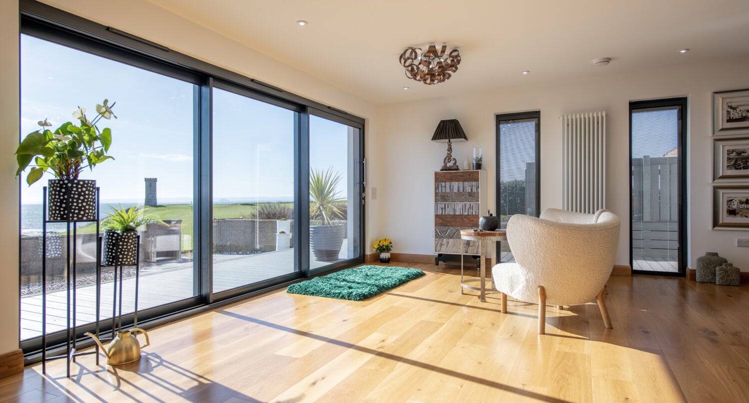 Bright, modern living room with large glass doors and bespoke glass solutions overlooking a grassy landscape and sea. Wooden floors, indoor plants, a cozy chair, and a small green rug decorate the sunlit space.