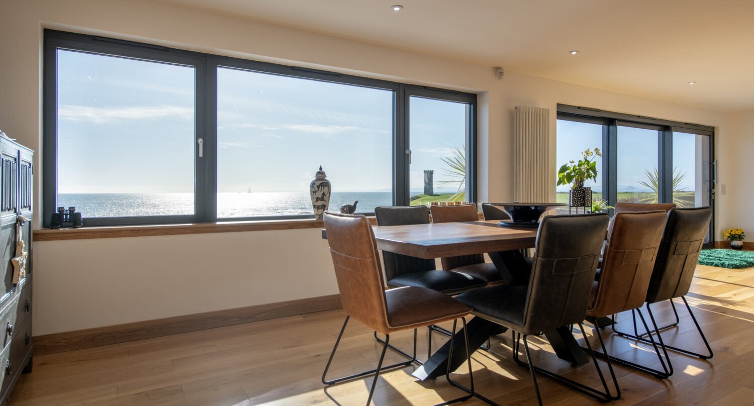 A modern dining room with a wooden table and leather chairs, large windows with bespoke glass solutions offering a sunny ocean view, wooden floors, and houseplants by the window.