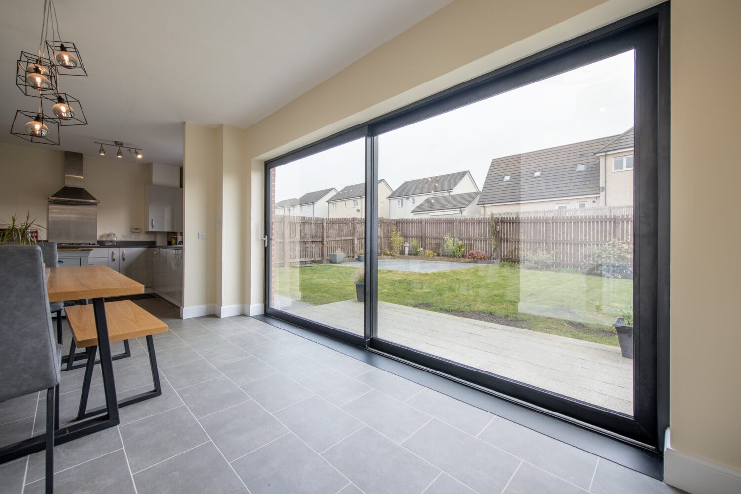 Modern dining area with bespoke glass solutions, including large sliding doors opening to a backyard garden. The room features tile flooring, wooden bench seating, contemporary pendant lights, and an adjacent view of the kitchen.