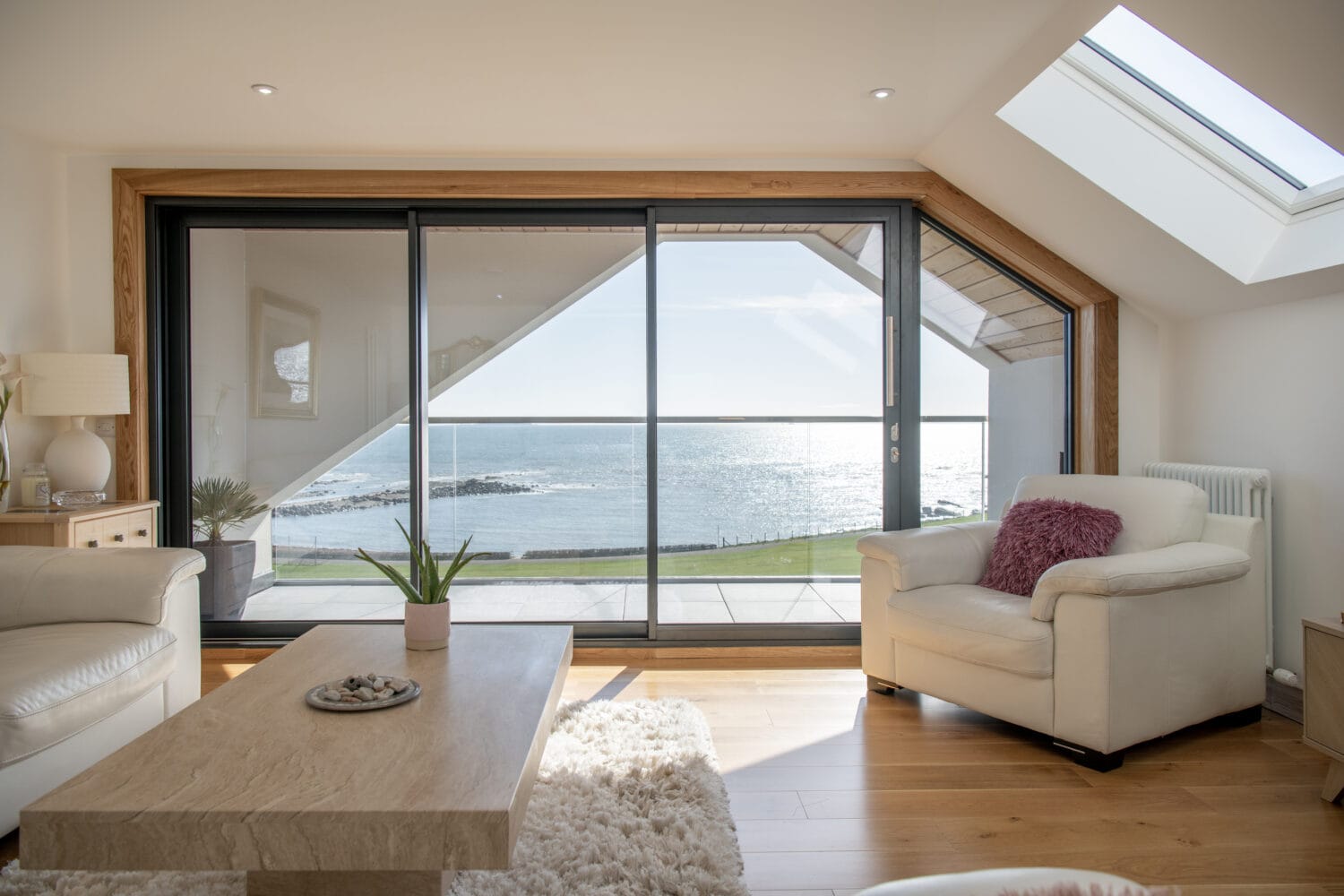 Modern living room with light wood floors, white armchairs, a coffee table, and sliding doors opening to a balcony with a scenic ocean view. Sunlight streams in through bespoke glass solutions, creating a bright and airy atmosphere.