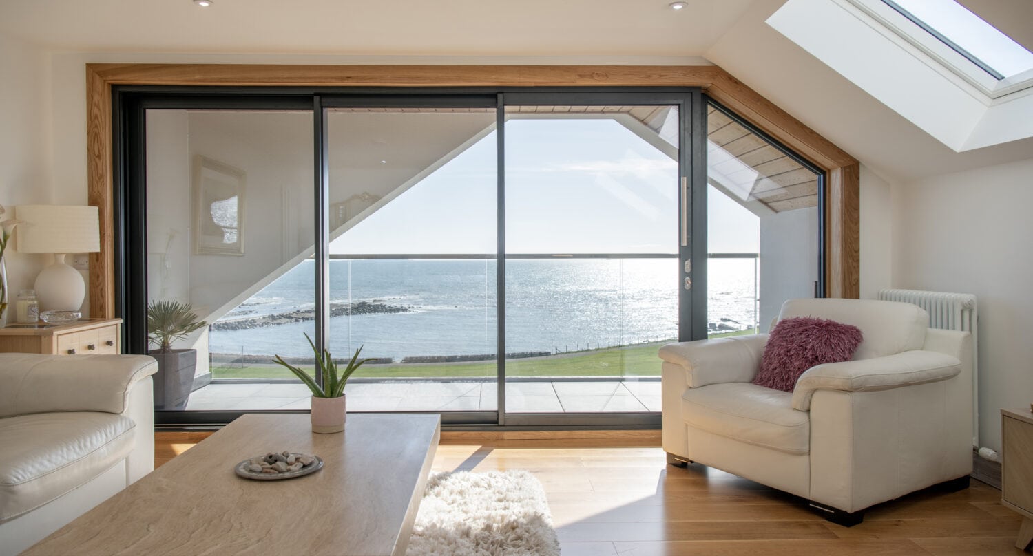 Modern living room with light wood floors, white armchairs, a coffee table, and sliding doors opening to a balcony with a scenic ocean view. Sunlight streams in through bespoke glass solutions, creating a bright and airy atmosphere.