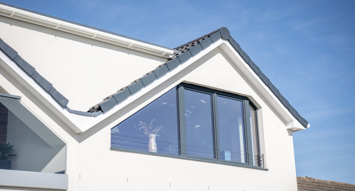 Modern white house with a pitched roof and large dark-framed window, reflecting blue sky. Bespoke glass solutions enhance the design, while a decorative vase with dried plants sits on the windowsill.