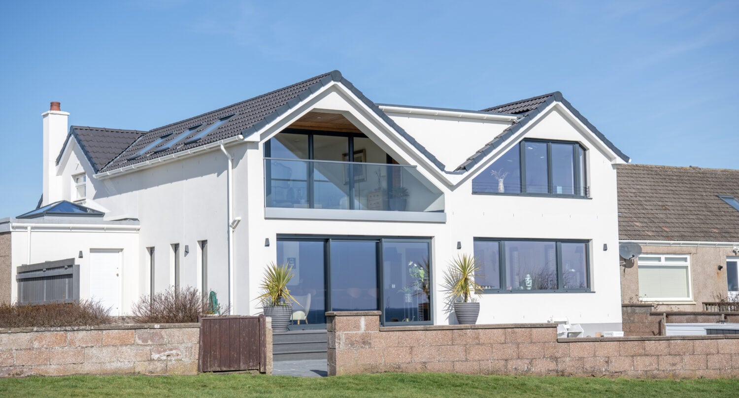 Modern two-story white house with large glass windows, sliding doors, and balcony, set against a clear blue sky. There is a stone wall, green lawn, and potted plants near the entrance.