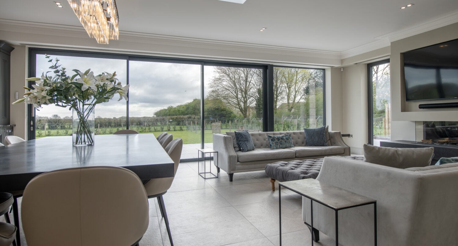 Modern open-plan living and dining area with large windows and sliding doors overlooking a green landscape, featuring a dining table with white chairs, elegant chandelier, sofas, and a wall-mounted TV.