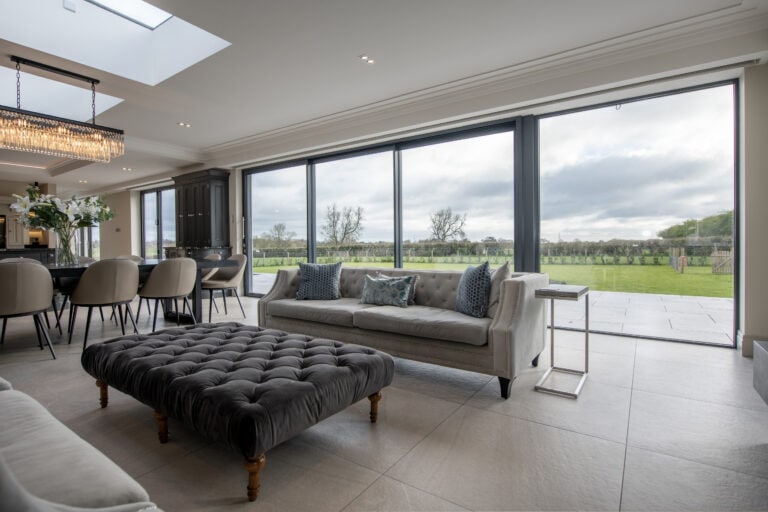 Modern open-plan living room with large floor-to-ceiling windows and sliding doors, light grey sofas, a dark ottoman, dining area with white chairs, and a view of a green lawn and trees outside under a cloudy sky.