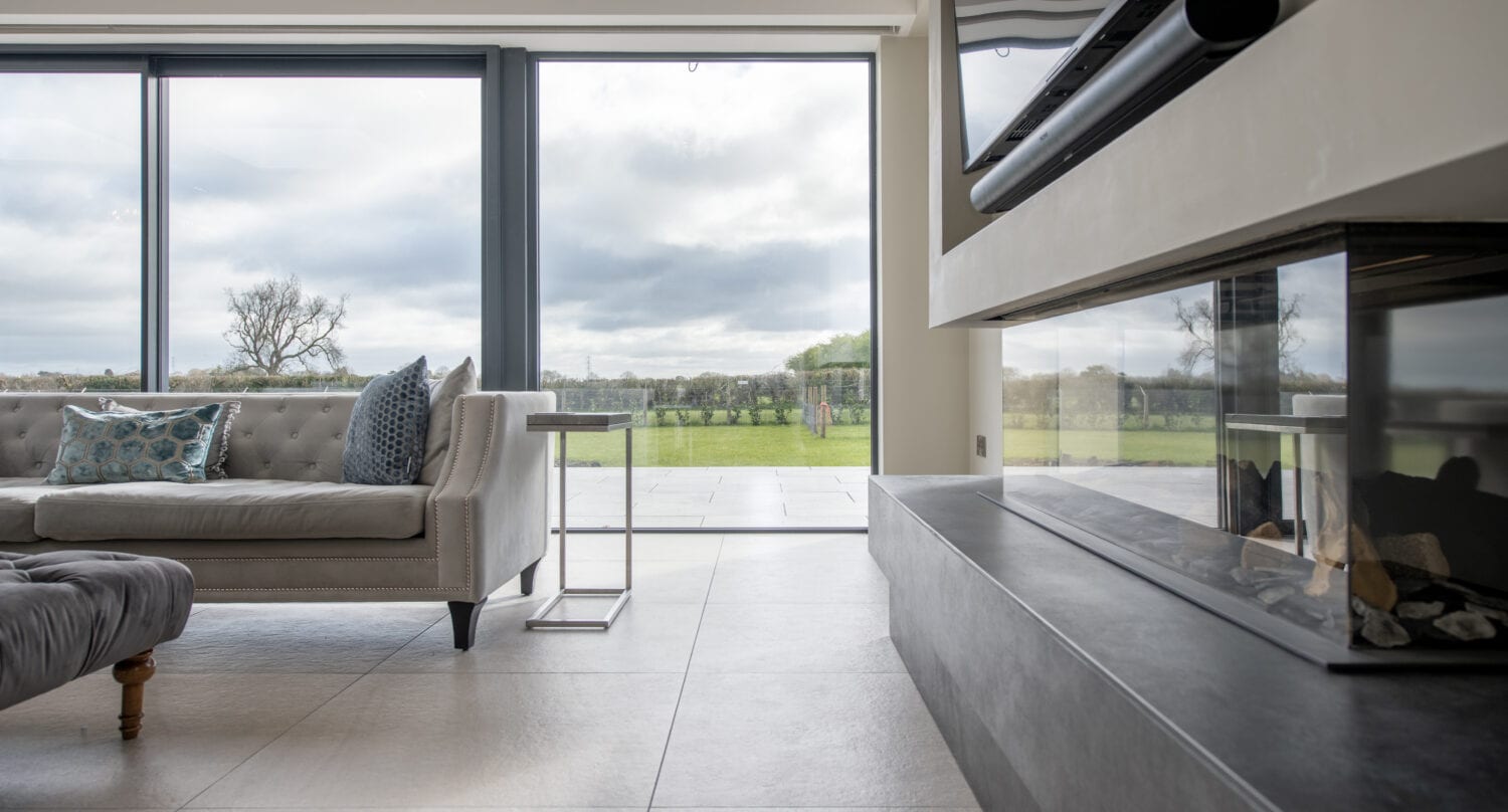 Modern living room with large floor-to-ceiling sliding doors, a gray sofa with patterned pillows, a glass side table, and a contemporary fireplace; outside, a green lawn and cloudy sky are visible.