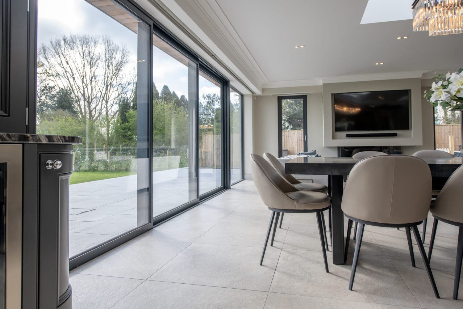 Modern dining room with beige chairs around a black table, large bespoke glass solutions like sliding doors open to a patio and green garden, wall-mounted TV, and plenty of natural light.