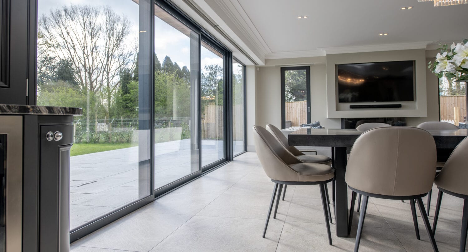 Modern dining room with beige chairs around a black table, large bespoke glass solutions like sliding doors open to a patio and green garden, wall-mounted TV, and plenty of natural light.