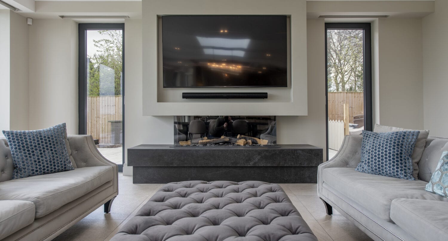 Modern living room with two light gray sofas, blue patterned cushions, a large tufted ottoman, sleek black fireplace with wall-mounted TV, tall windows, and bespoke glass solutions like glass roofing that enhance the space with abundant natural light.