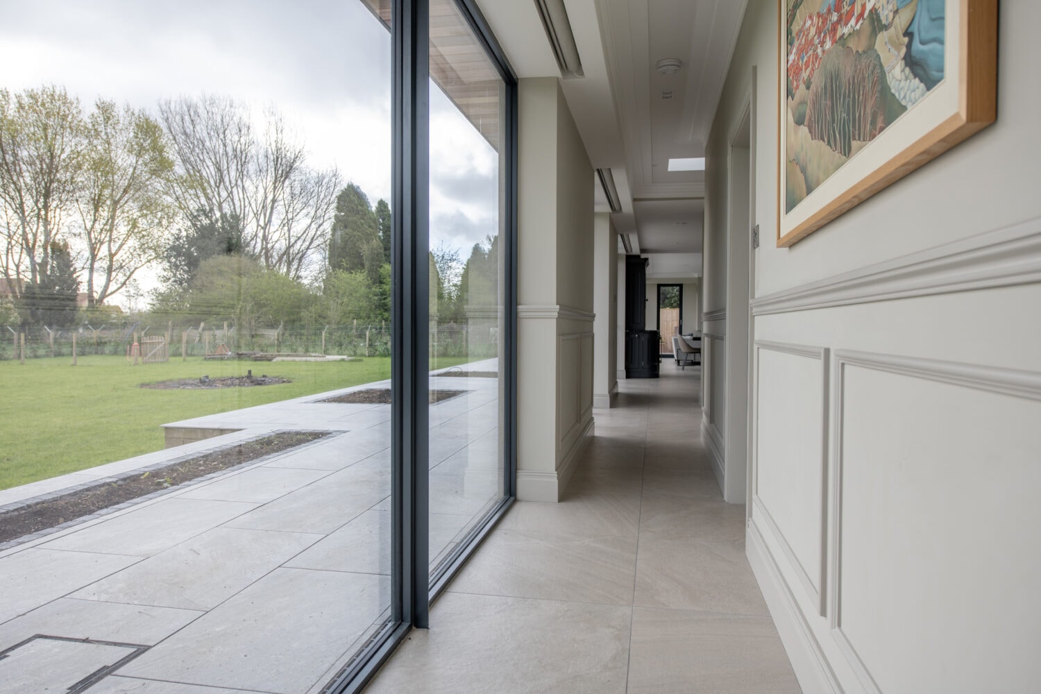 A bright hallway with large bifold doors on the left, overlooking a green garden. The right side features white paneled walls and a colorful framed painting, creating an inviting space enhanced by bespoke glass solutions.