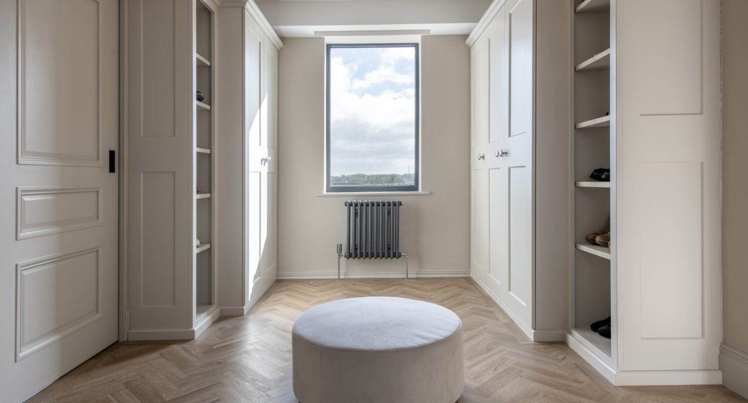 A bright, modern walk-in closet with built-in white cabinets on both sides, a round upholstered ottoman in the center, herringbone wood flooring, and a window with glass roofing above for stunning views of the sky and trees.