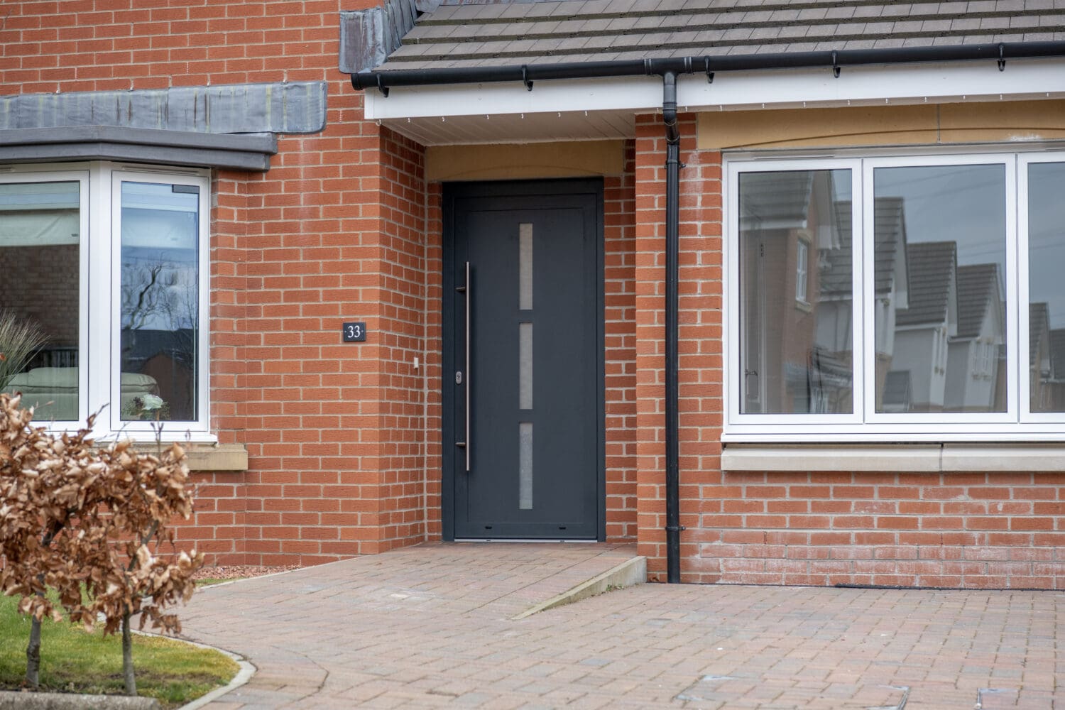 Modern brick house entrance with a dark gray front door, large windows featuring bespoke glass solutions, and a paved walkway. House number 33 is displayed next to the door, and a small shrub is visible in the foreground.