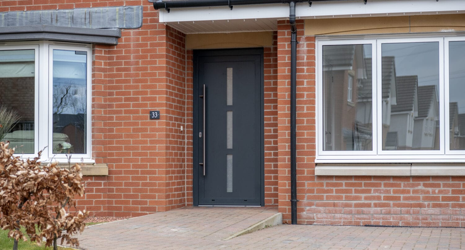 Modern brick house entrance with a dark gray front door, large windows featuring bespoke glass solutions, and a paved walkway. House number 33 is displayed next to the door, and a small shrub is visible in the foreground.