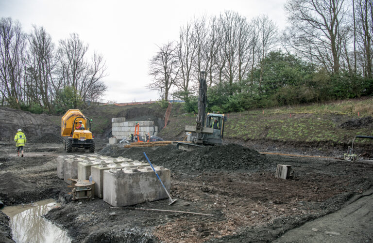 A construction site with heavy machinery, large concrete blocks, and two workers in safety gear installing sliding doors. Trees and grass fill the background, while the muddy, uneven ground stretches across the foreground.