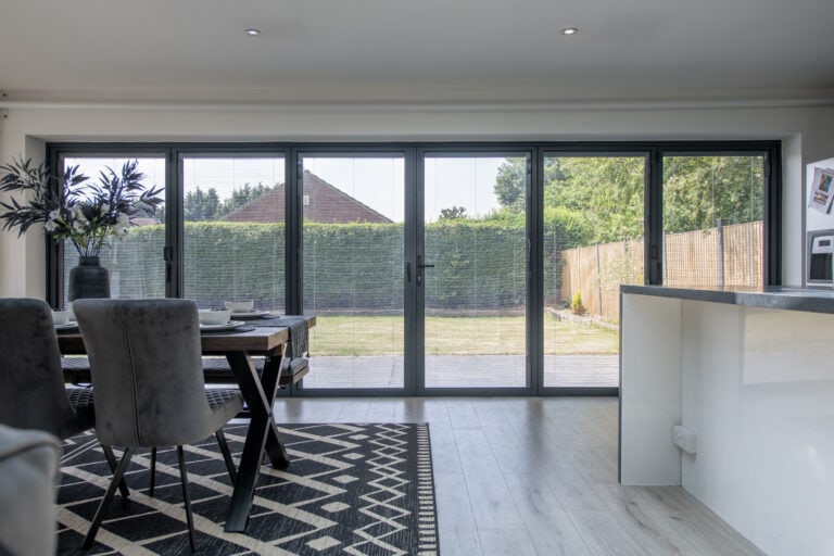 A modern dining area with a table and chairs, a patterned rug, and large bespoke glass solutions folding doors opening to a garden with a lawn and tall green hedges outside.