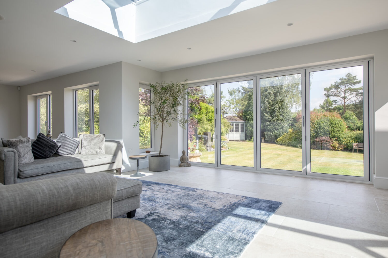 A bright, modern living room with large sliding doors and windows overlooking a lush garden. There’s a gray sectional sofa, cushions, a small table, a rug, and a potted plant. Sunlight fills the space, highlighting bespoke glass solutions.