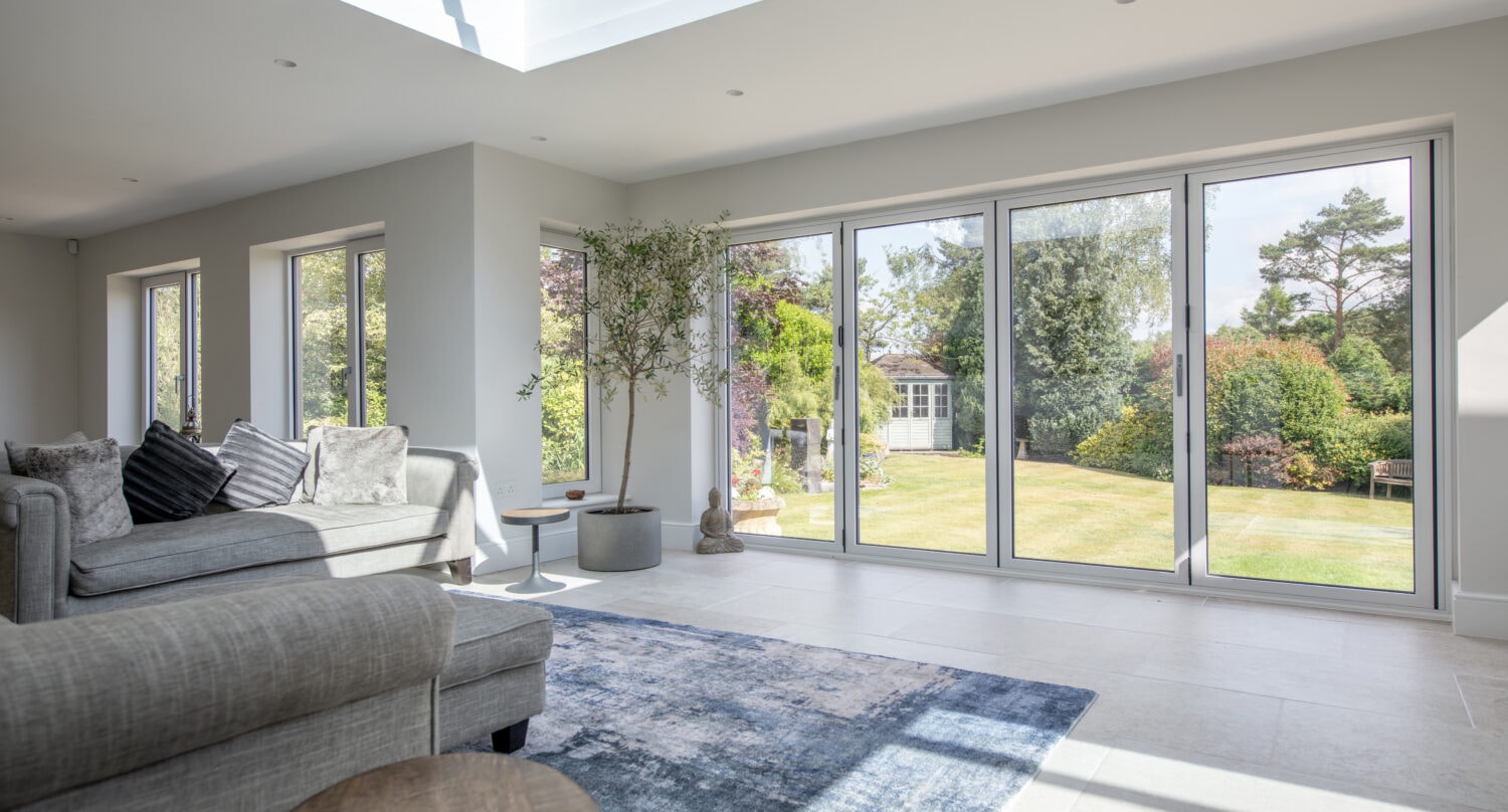 A bright, modern living room with large sliding doors and windows overlooking a lush garden. There’s a gray sectional sofa, cushions, a small table, a rug, and a potted plant. Sunlight fills the space, highlighting bespoke glass solutions.