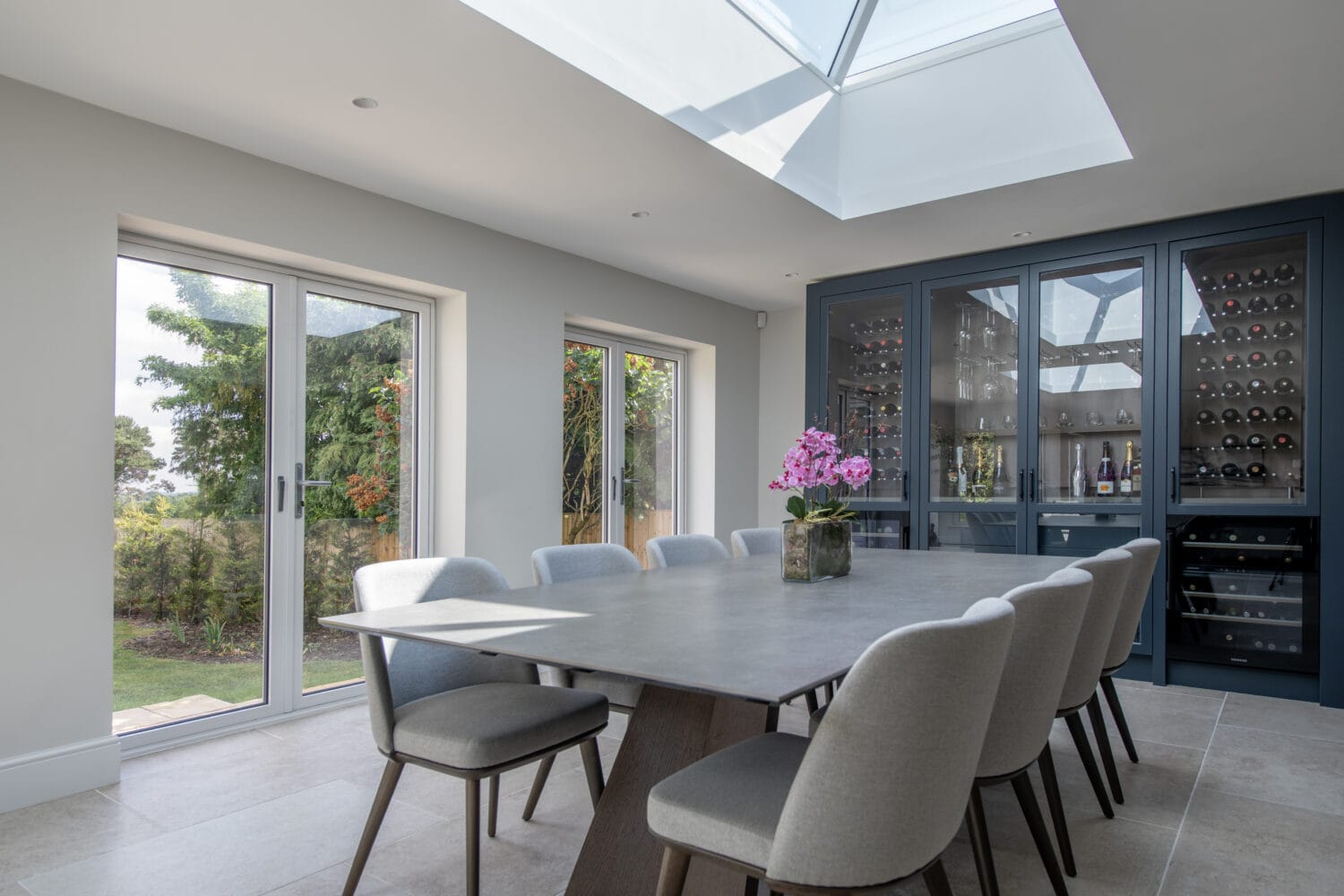 A modern dining room with a large table, eight gray chairs, a skylight, bespoke glass solutions like sliding doors to the garden, and a built-in dark cabinet displaying wine bottles and glassware. A pink orchid adorns the table.