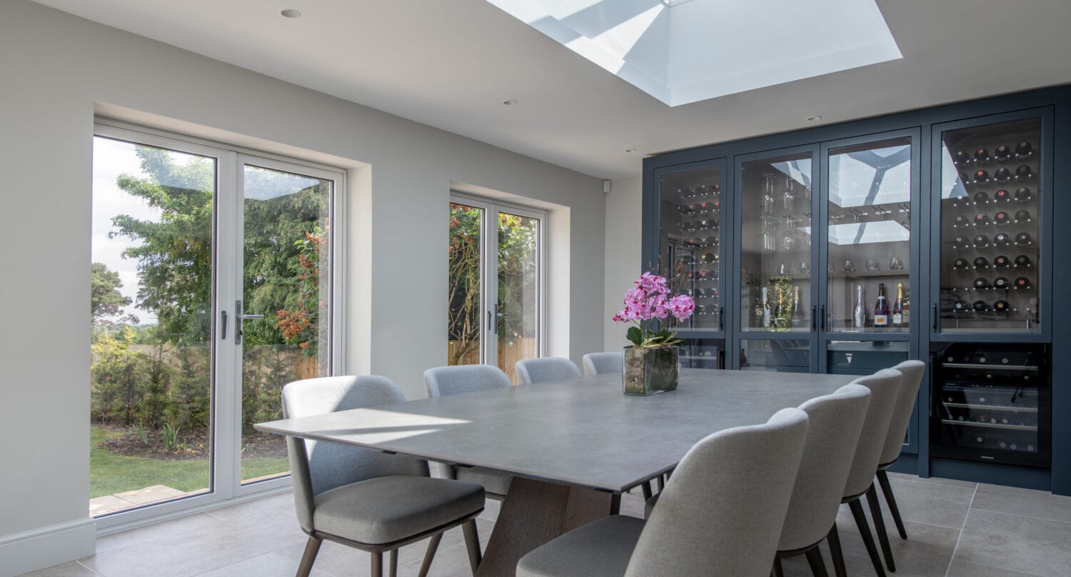 A modern dining room with a large table, eight gray chairs, a skylight, bespoke glass solutions like sliding doors to the garden, and a built-in dark cabinet displaying wine bottles and glassware. A pink orchid adorns the table.