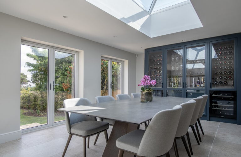 A modern dining room with a large table, eight gray chairs, a skylight, bespoke glass solutions like sliding doors to the garden, and a built-in dark cabinet displaying wine bottles and glassware. A pink orchid adorns the table.