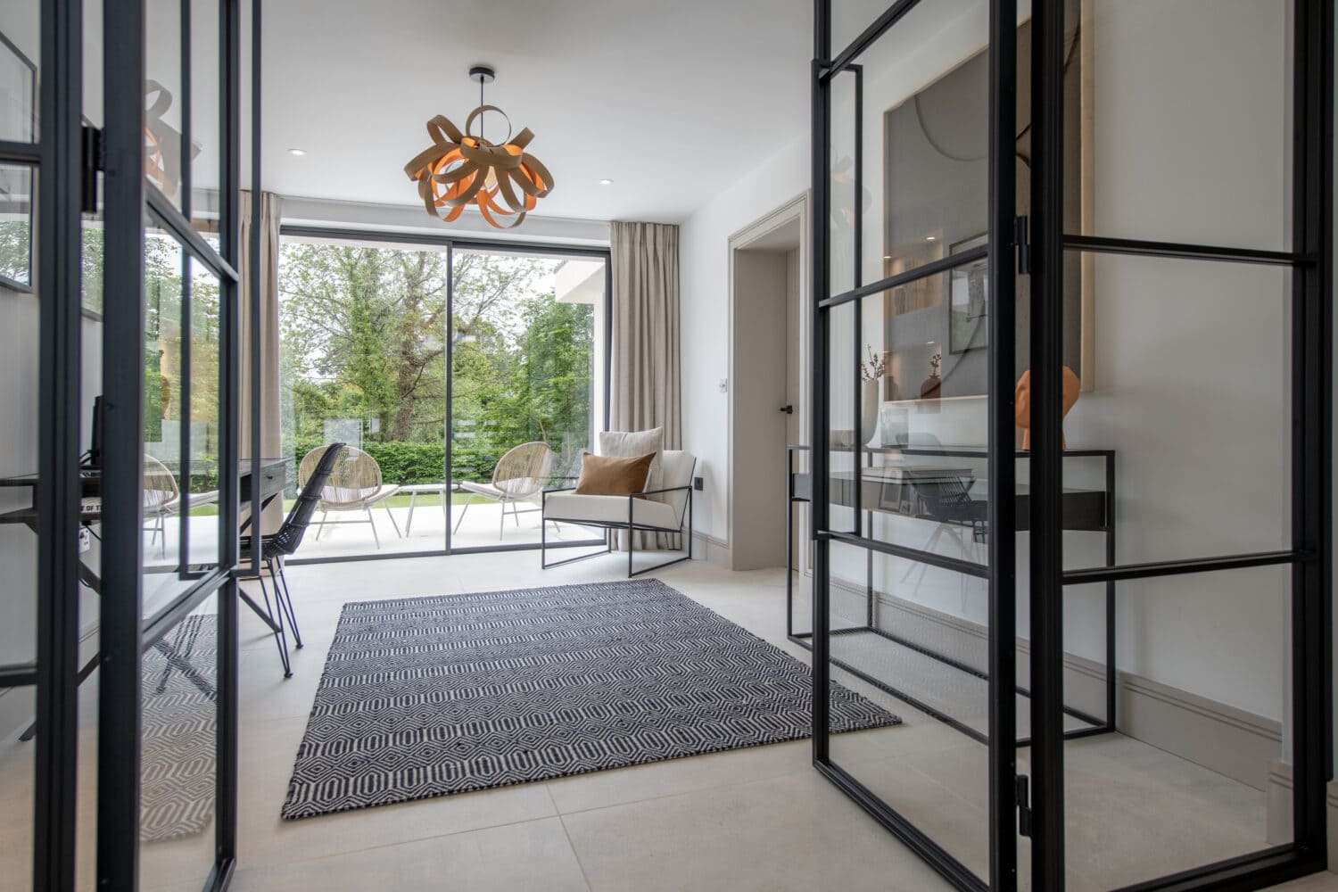 A modern, bright living space with bespoke glass solutions, geometric rug, contemporary chairs, a decorative light fixture, and large windows offering a view of a green garden outside.