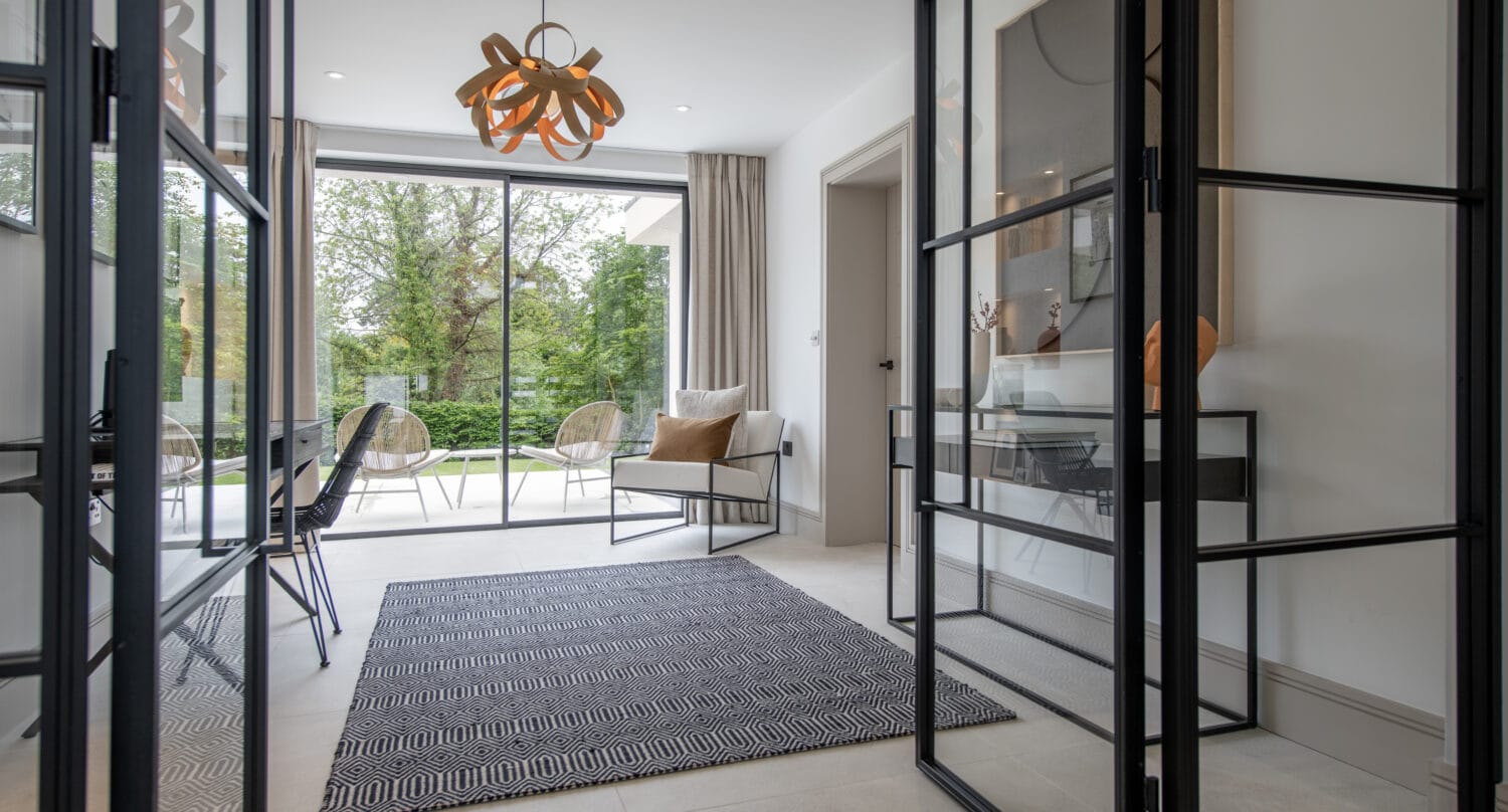 A modern, bright living space with bespoke glass solutions, geometric rug, contemporary chairs, a decorative light fixture, and large windows offering a view of a green garden outside.
