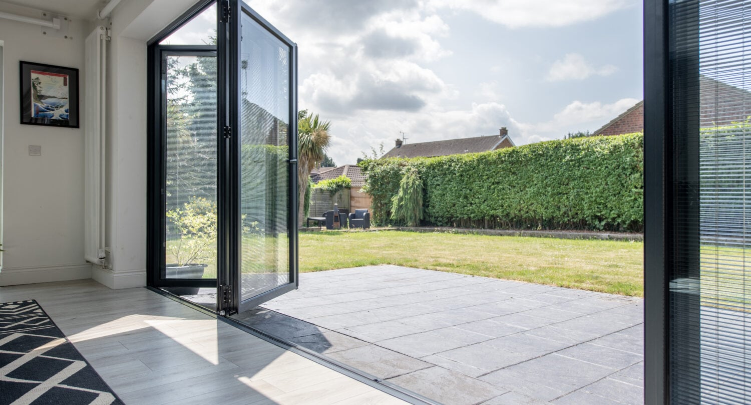 A set of bespoke glass folding doors opens from a modern living room onto a stone patio and a grassy backyard, bordered by a tall green hedge under a partly cloudy sky.