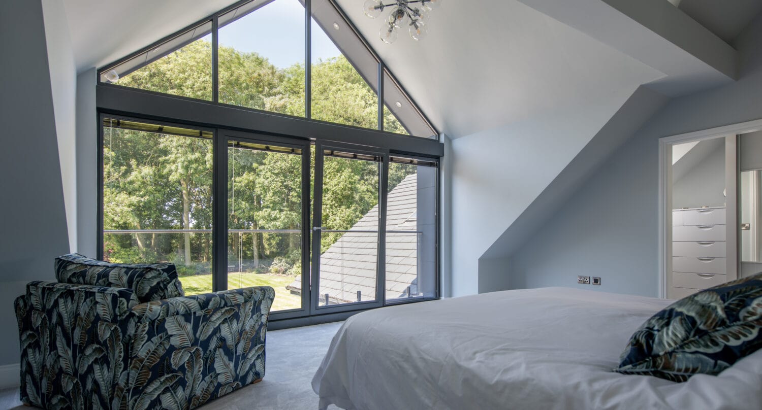 Modern bedroom with sloped ceiling, large floor-to-ceiling windows, and sliding doors that open to a view of trees. The room features a patterned armchair, a bed with white bedding, and a contemporary chandelier.