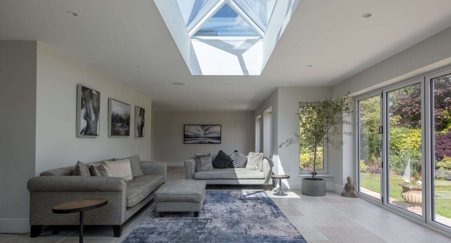 A modern living room with two gray sofas, a blue area rug, wall art, and large bifold doors opening to a garden. A skylight above lets in natural light, casting shadows on the floor—showcasing bespoke glass solutions.