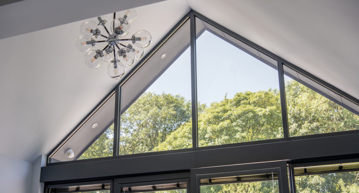 Large triangular window with black frames lets in natural light, revealing green trees outside. Glass roofing enhances the airy feel, while a modern chandelier with multiple bulbs hangs from the white, vaulted ceiling.