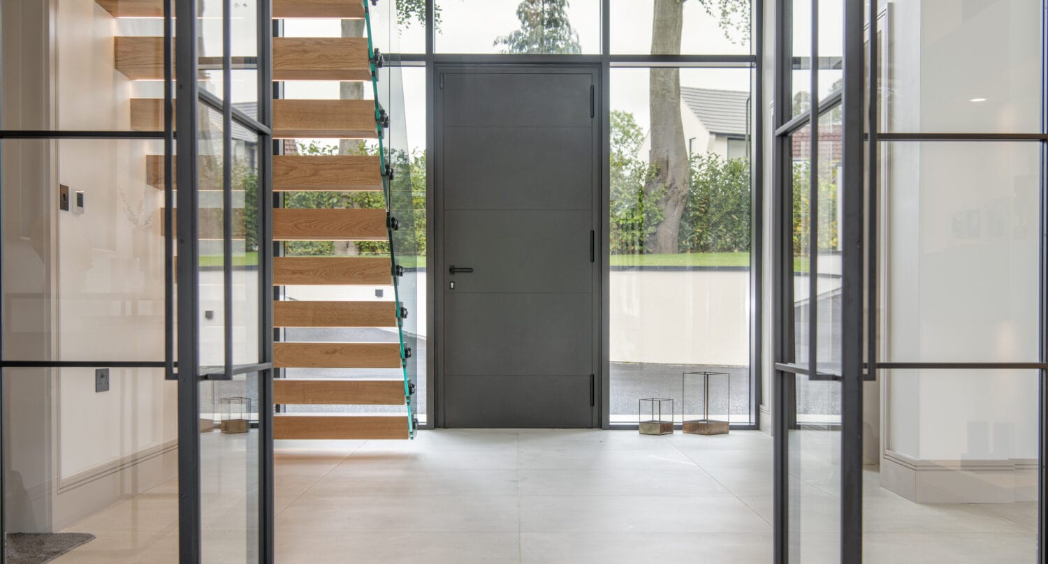 A modern, bright entryway featuring a wooden floating staircase with glass railings, large windows, a sleek black front door, and minimalist decor, allowing natural light to fill the space through stylish front doors.