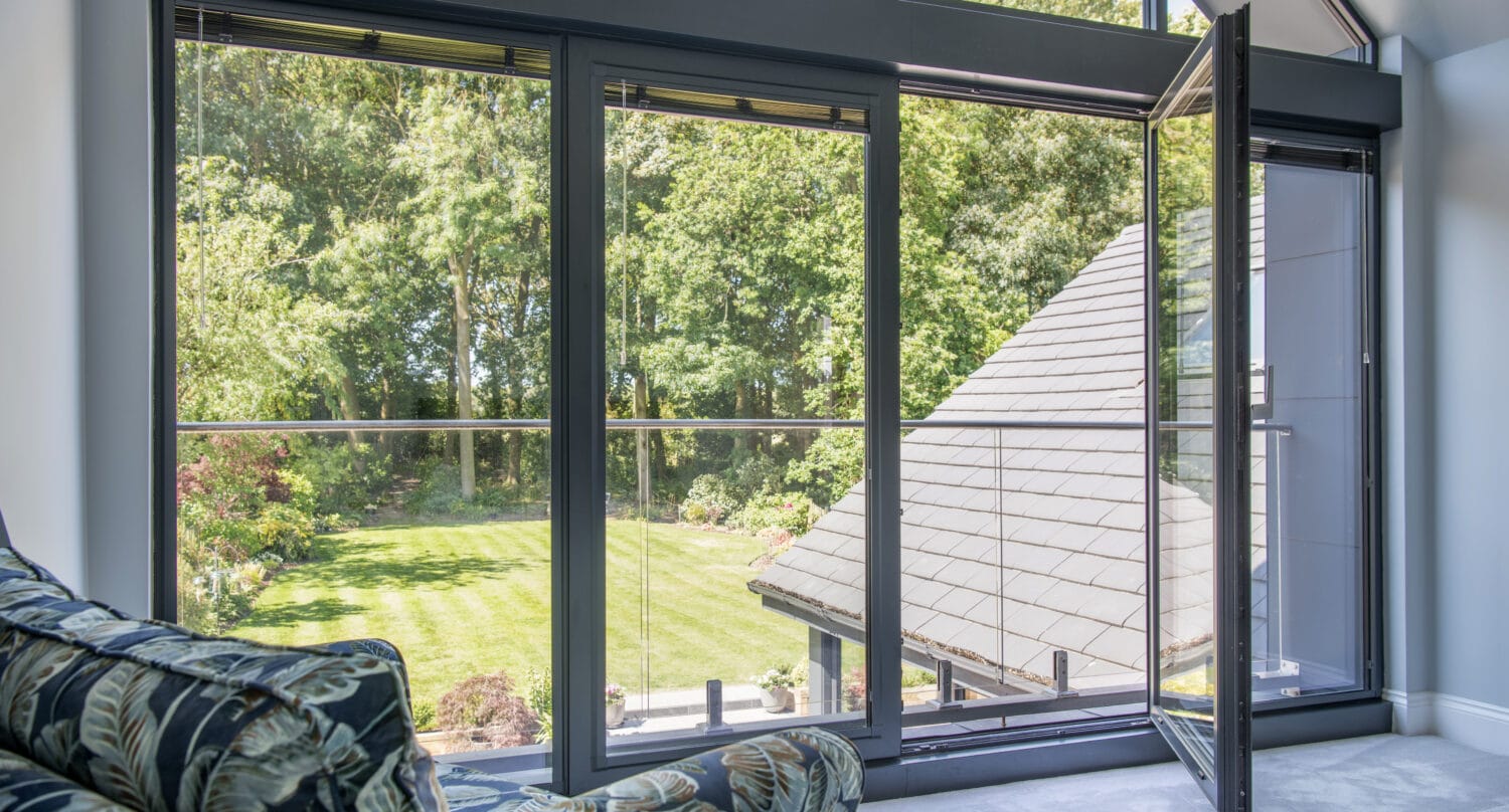 A modern bifold door opens to a balcony overlooking a green, well-kept lawn and trees. Sunlight streams in, and part of a patterned sofa is visible in the foreground.