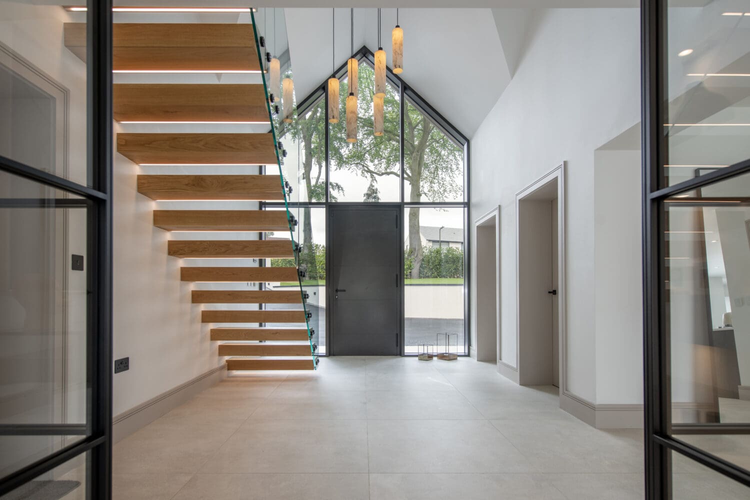 Modern entryway with floating wooden stairs on the left, tall glass windows, bespoke glass solutions, a black front door, pendant lights hanging from the ceiling, and two doorways on the right. The space is bright and minimalistic.