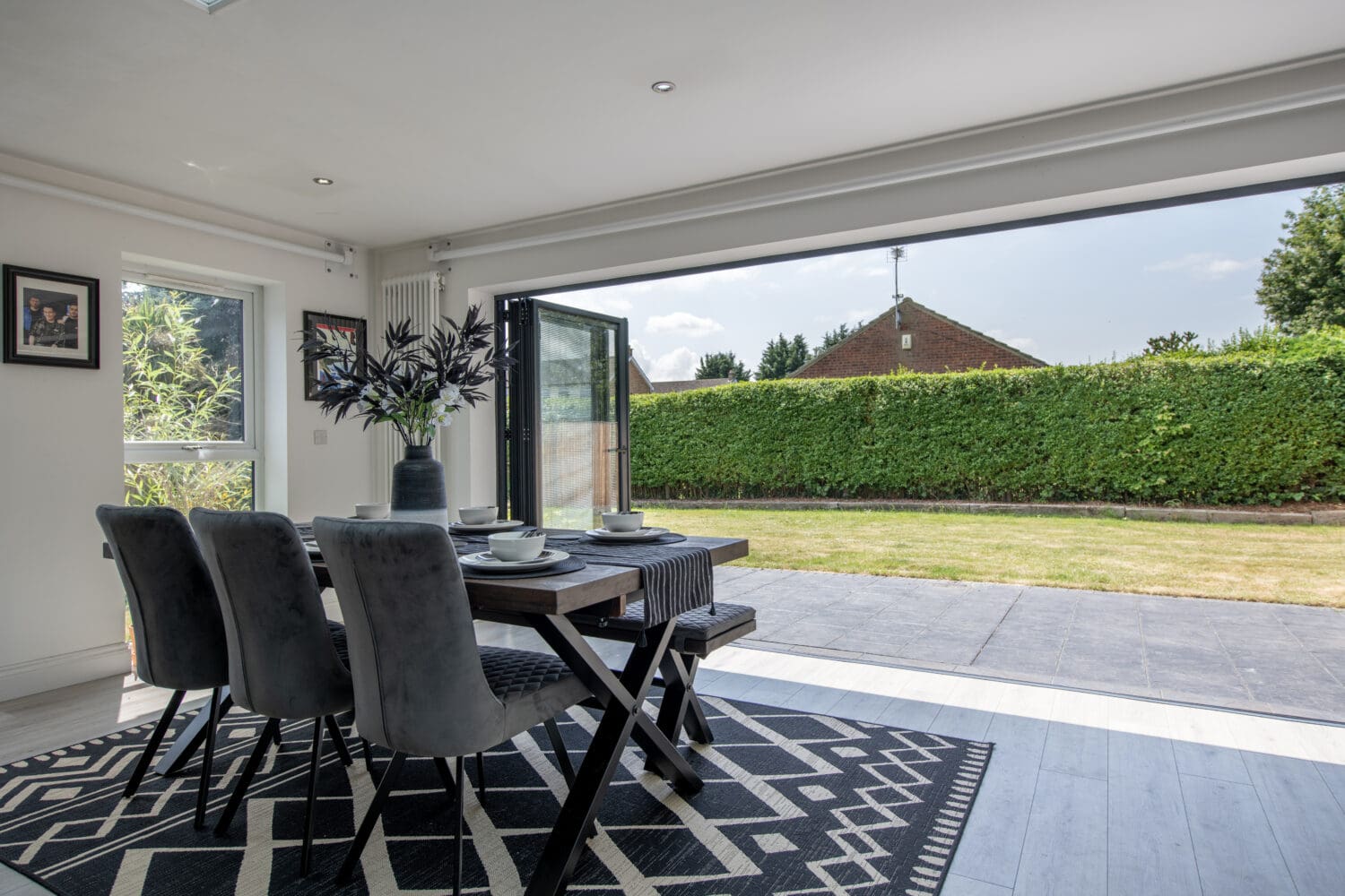 A modern dining area with a black table and four gray chairs sits on a patterned rug beside wide open sliding doors, overlooking a green garden and a hedged lawn under a sunny sky.