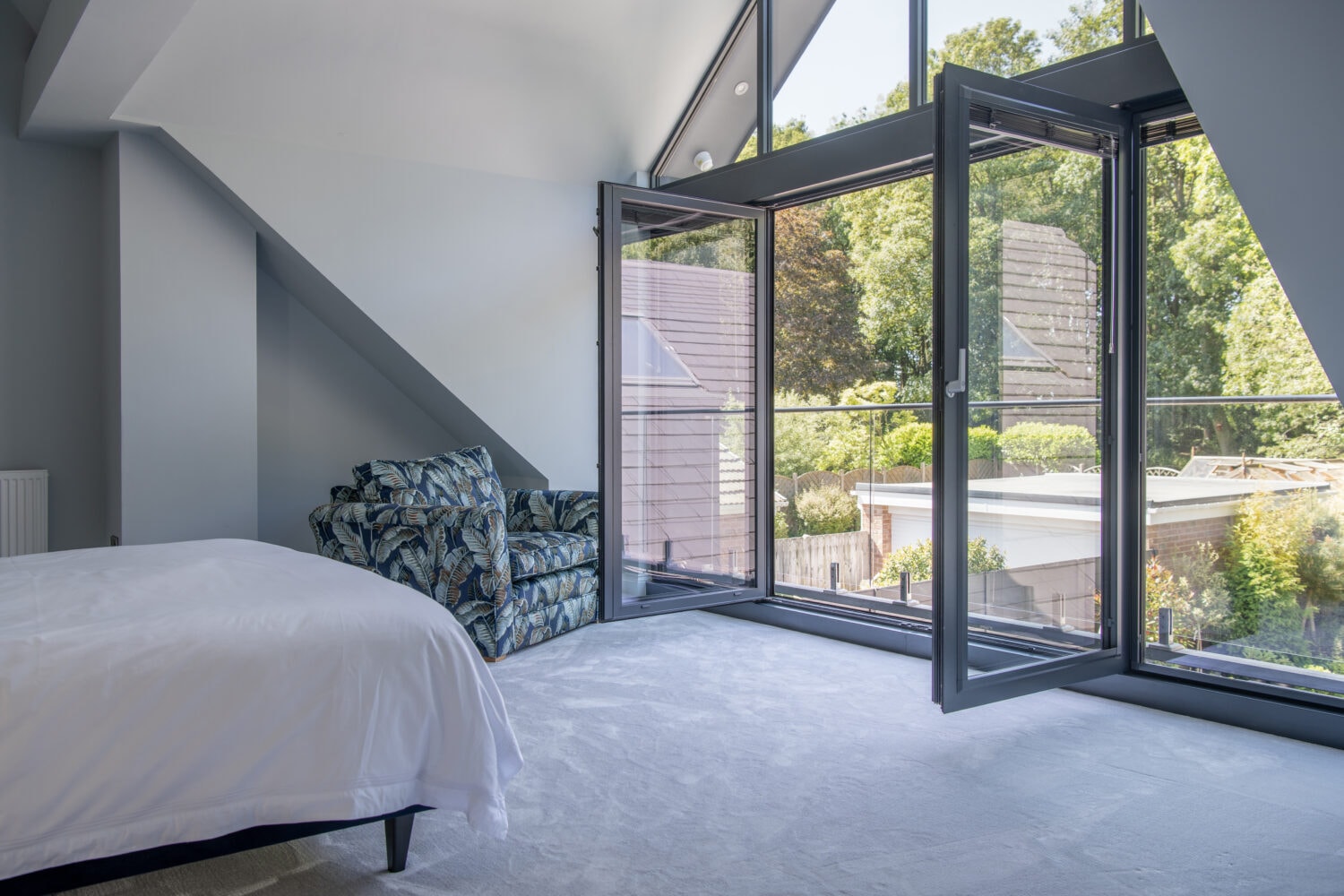 A bright, modern bedroom features bespoke glass solutions with large sliding doors open to a balcony, a white bed, a patterned armchair, light carpet, and trees visible outside.