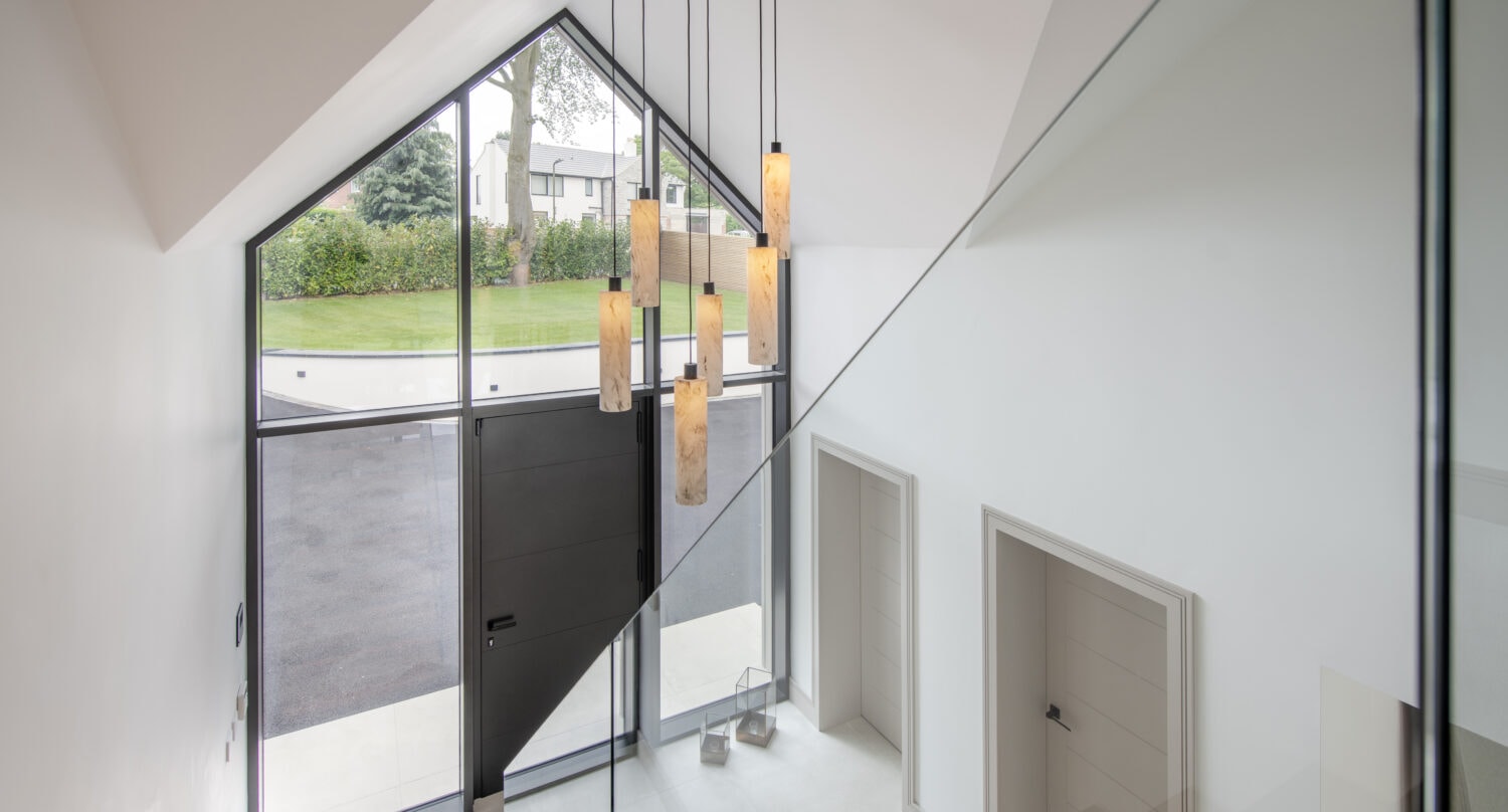 Modern entryway with tall bespoke glass front doors, high ceilings, large triangular window, cluster of pendant lights, white walls, tiled floor, and two closed doors beneath a glass staircase. Lawn and trees visible outside.