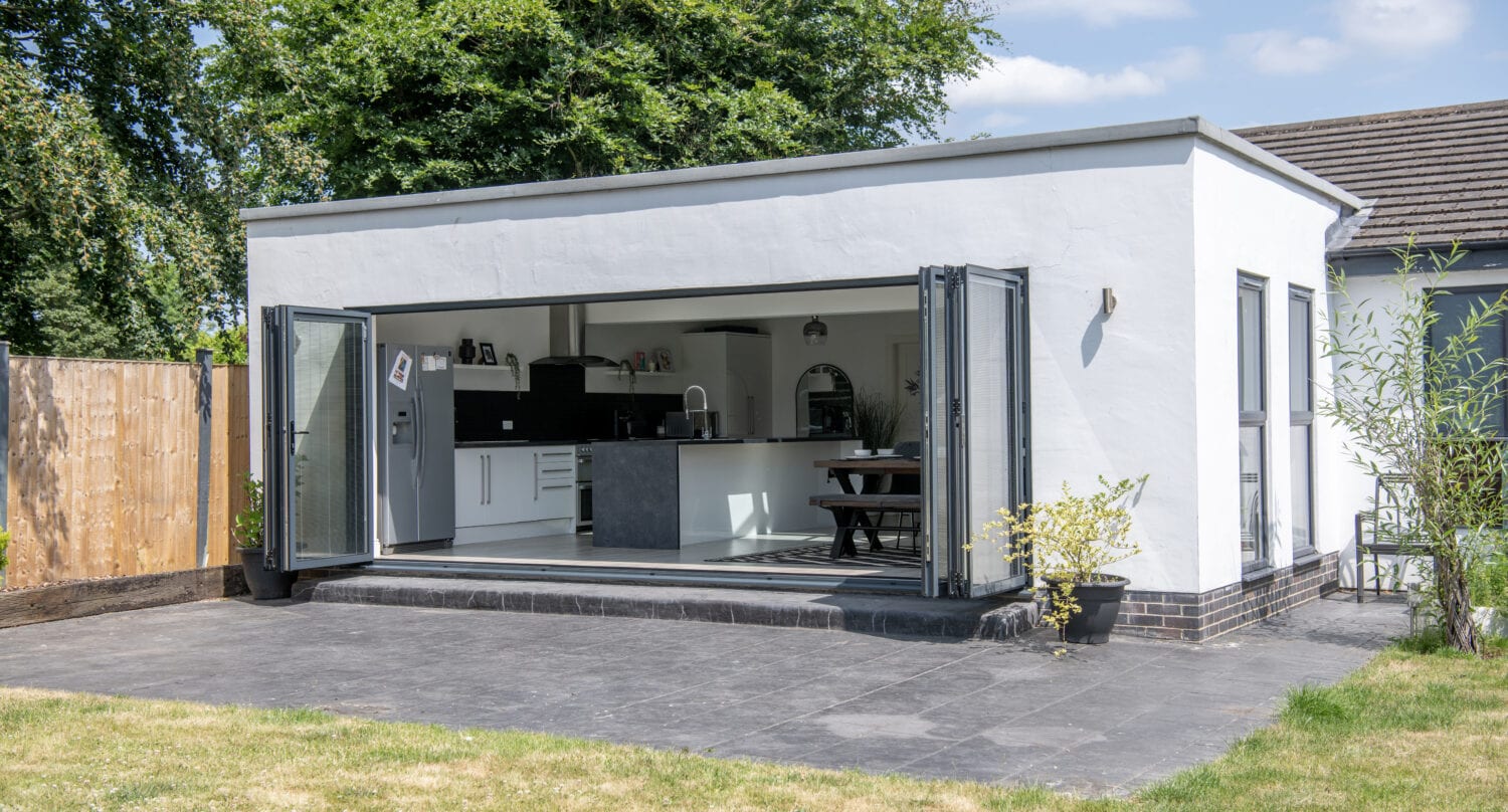 A modern white extension with large bifold doors opens onto a patio, revealing a stylish kitchen and dining area. The space is surrounded by a grassy lawn and some plants, with trees visible in the background.