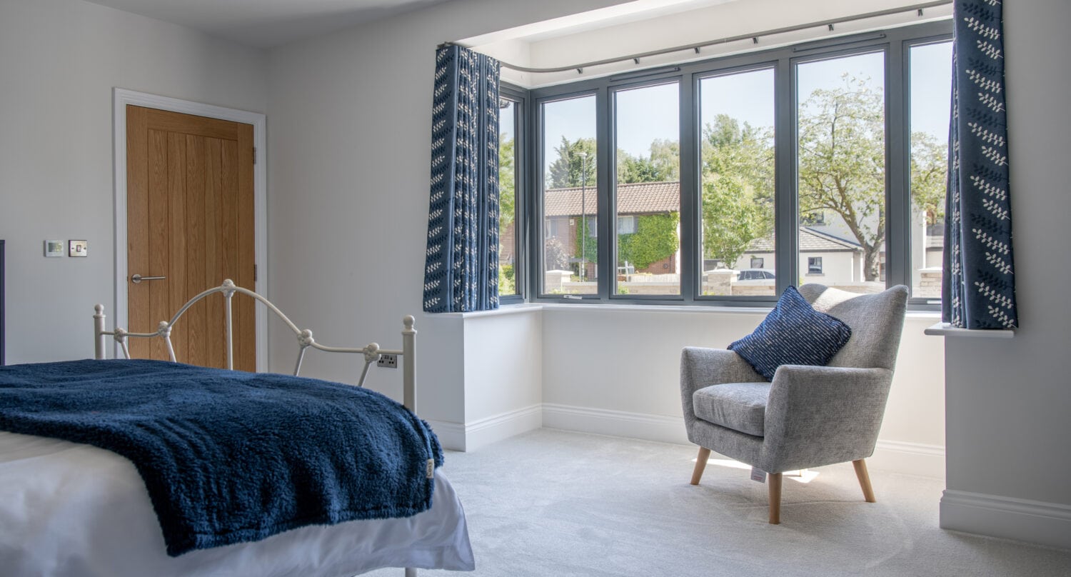 Bright bedroom with a large bay window, blue curtains, and a cozy grey armchair with blue cushions. Sunlight streams in through sliding doors, highlighting the white bed’s blue throw and the light carpet amid minimalist decor.