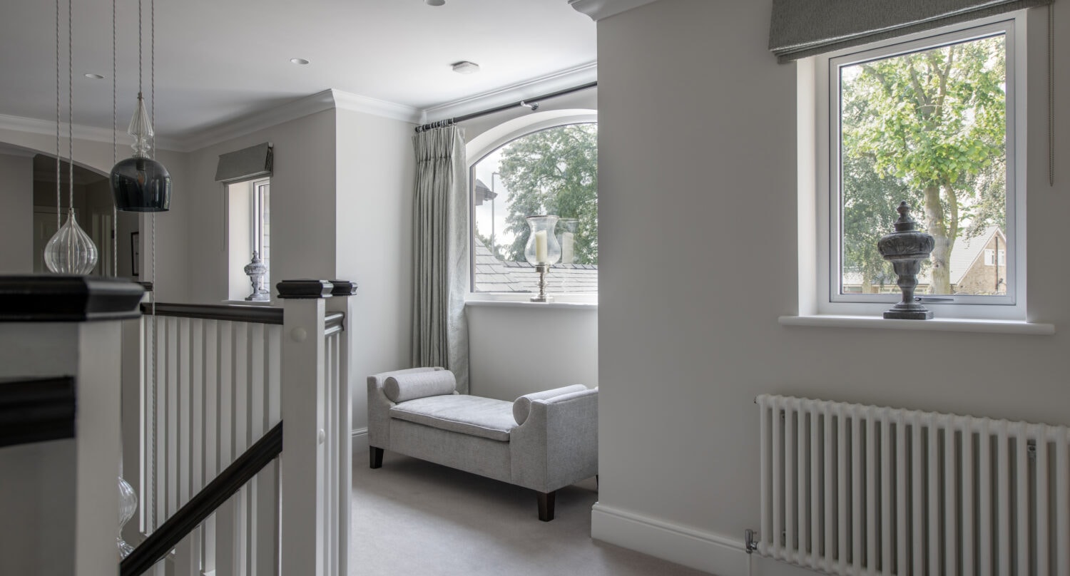 A bright, modern hallway with light gray walls, a large window, bespoke glass solutions, and a cushioned bench under an arched window. Sunlight streams in, highlighting minimalist decor and a white radiator beneath a second window.