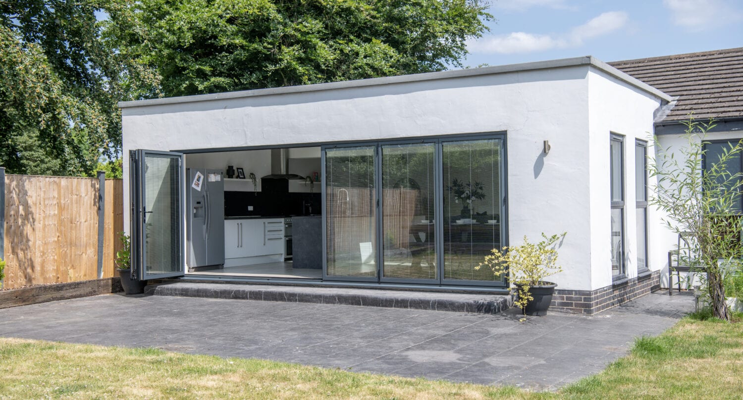 A modern white house extension with glass roofing and large sliding doors opens onto a stone patio and green lawn; the interior kitchen is visible, and potted plants sit outside.
