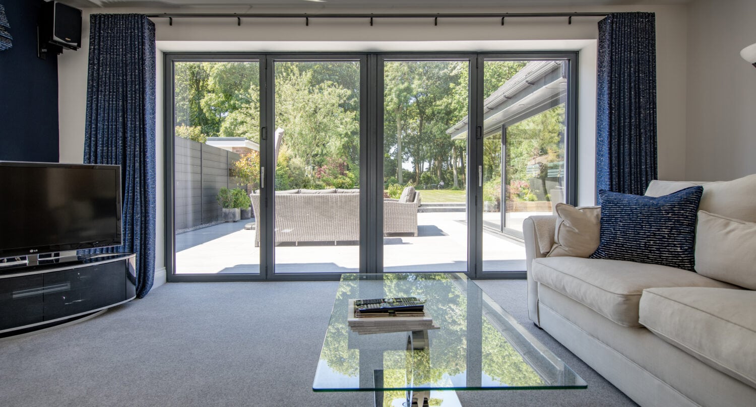 A modern living room with a beige sofa, glass coffee table, and TV, featuring bespoke glass solutions with large sliding doors that open to a sunny patio and green garden. Blue curtains frame the doors as natural light fills the space.