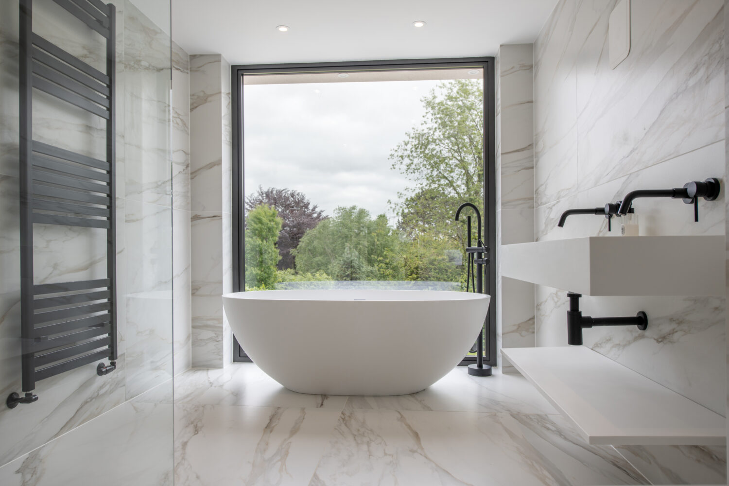 Modern bathroom with a freestanding white bathtub in front of large sliding doors overlooking trees. The room features marble walls and floors, a wall-mounted sink, black fixtures, and a heated towel rail.