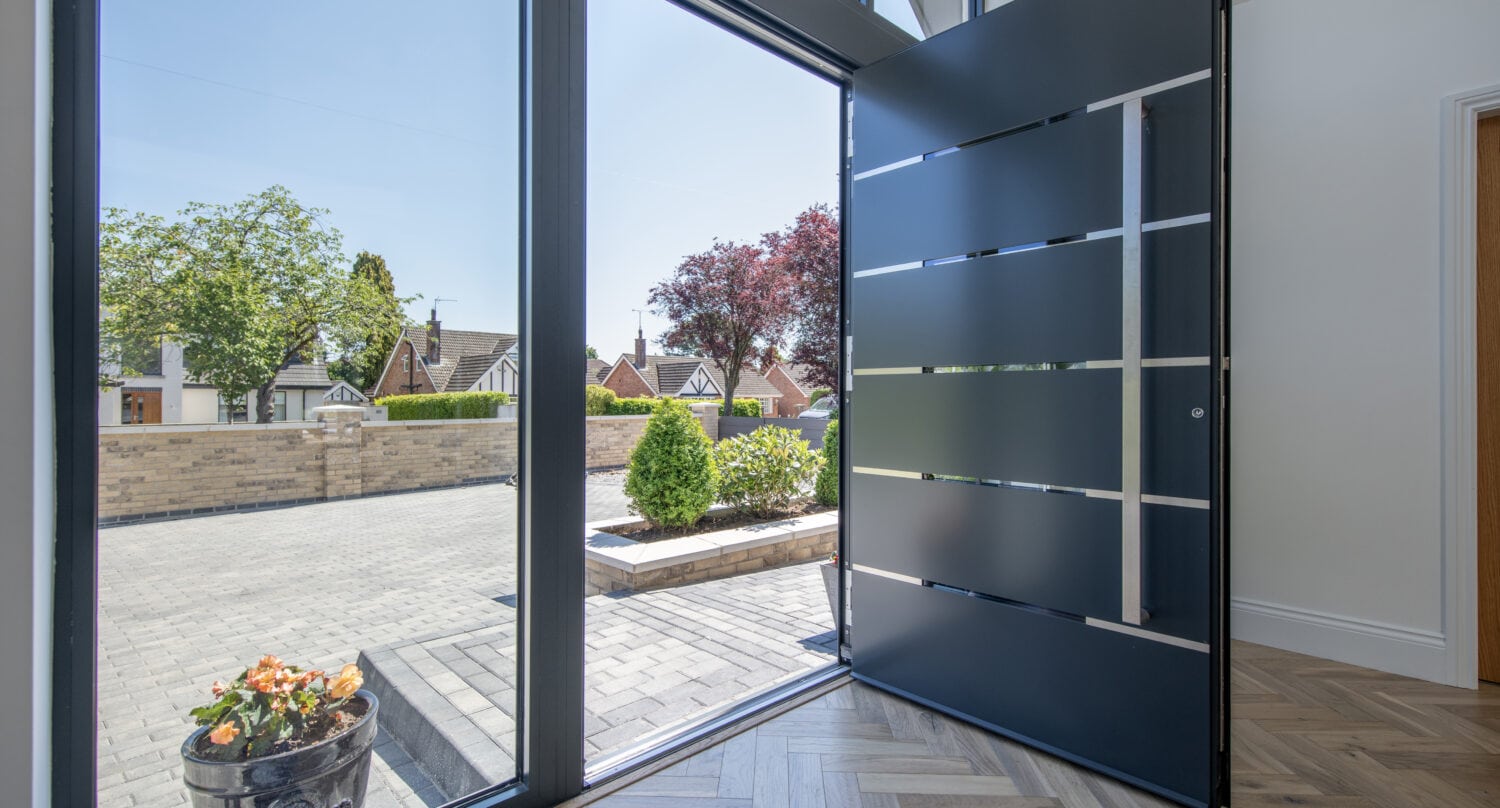 A modern home entrance with a large, open black front door featuring horizontal metal accents and bespoke glass solutions, letting in sunlight. Outside, a paved driveway, green shrubs, and trees are visible on a bright day.