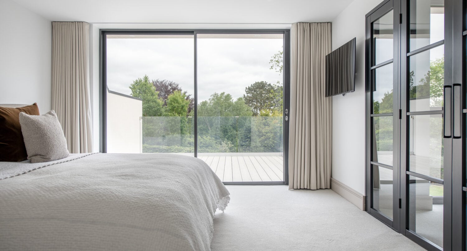 Modern bedroom with a neatly made bed, large bespoke glass doors leading to a balcony with tree views, beige curtains, a wall-mounted TV, and glass-paneled double doors on the right. The room is bright and minimalistic.