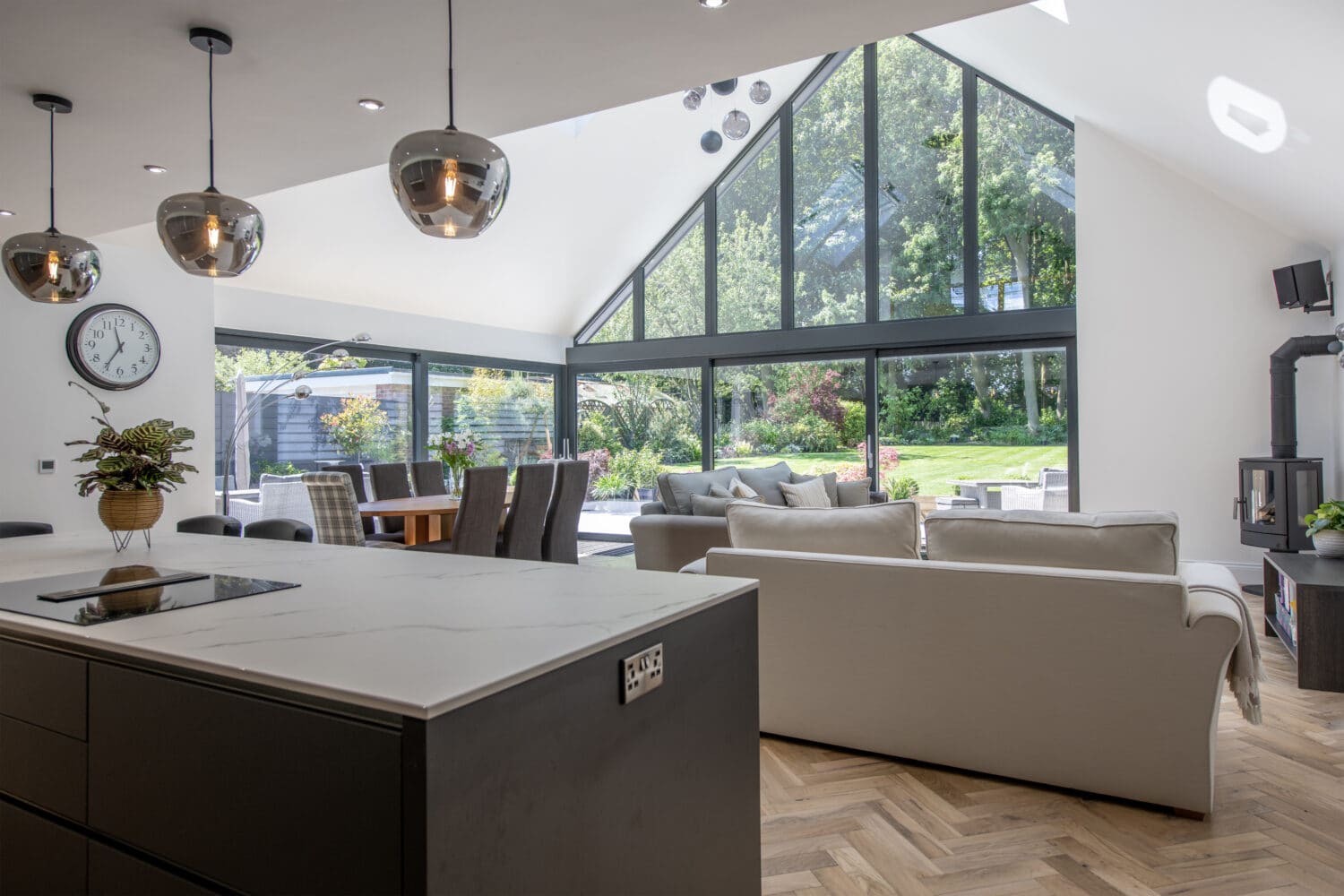 Modern open-plan living space with a kitchen island, dining table, and two sofas. Floor-to-ceiling windows by bespoke glass solutions offer lush garden views, filling the room with natural light.