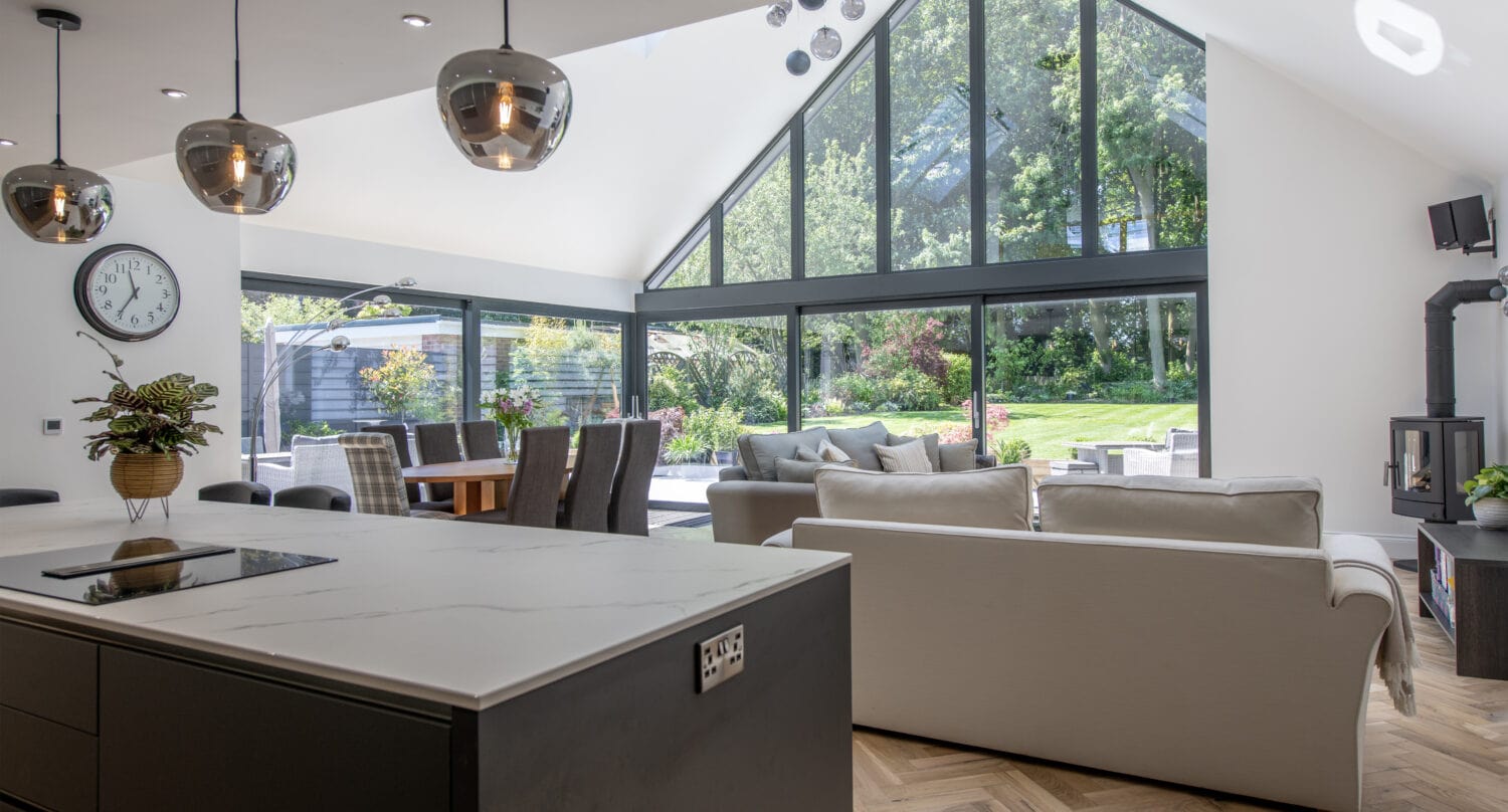 Modern open-plan living space with a kitchen island, dining table, and two sofas. Floor-to-ceiling windows by bespoke glass solutions offer lush garden views, filling the room with natural light.