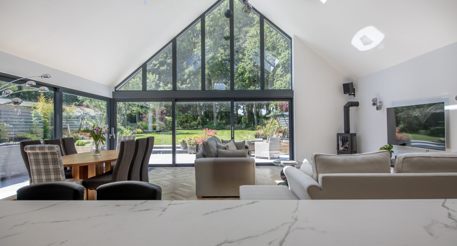 Modern, spacious living room with high vaulted ceiling, large bifold doors and skylights, neutral furniture, dining area with chairs, a garden view, and natural light filling the space.