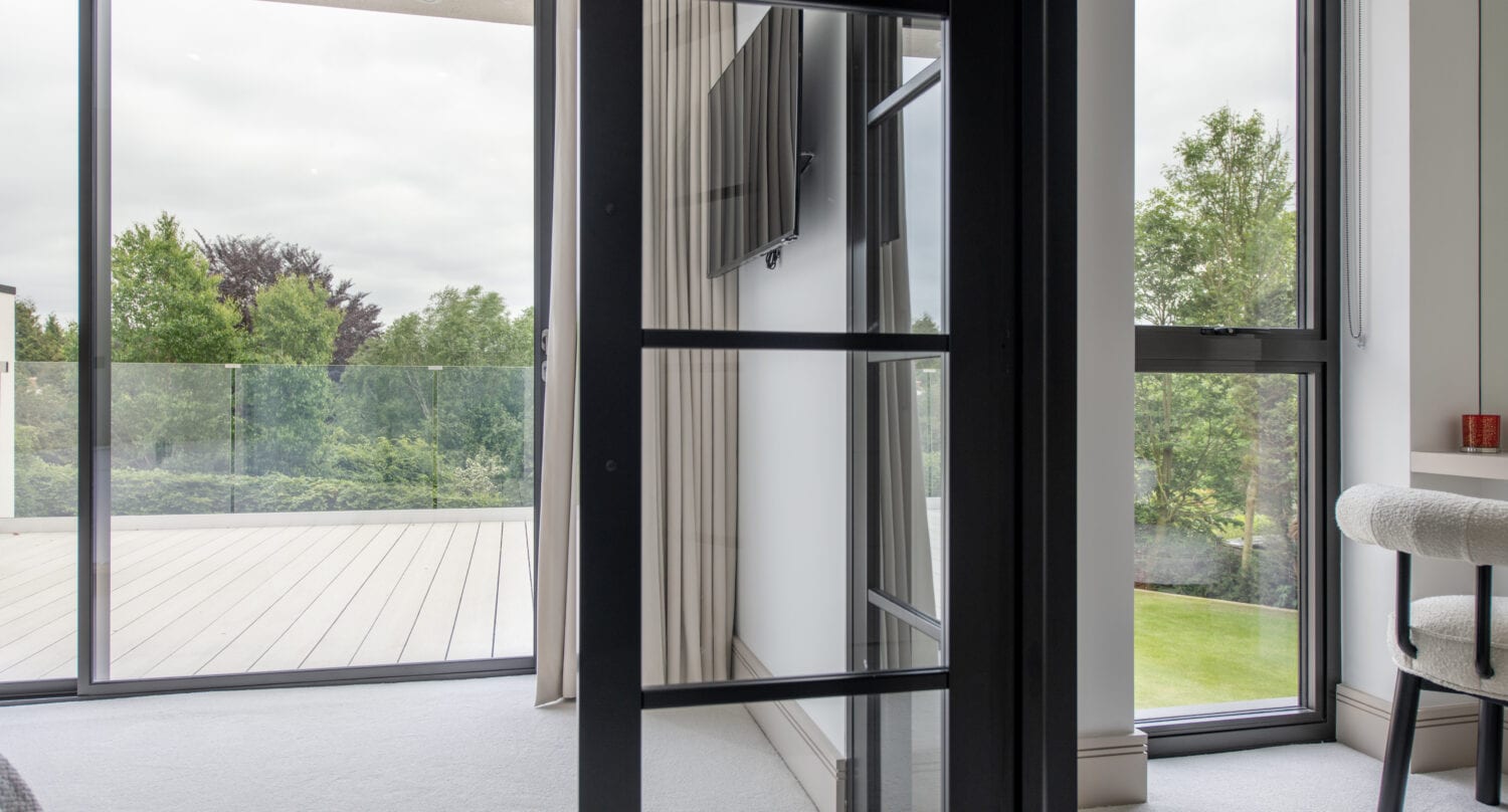 Modern room with large sliding doors and windows overlooking a green outdoor landscape, featuring a wall-mounted TV, a chair by a small table, and an open patio with a glass railing.