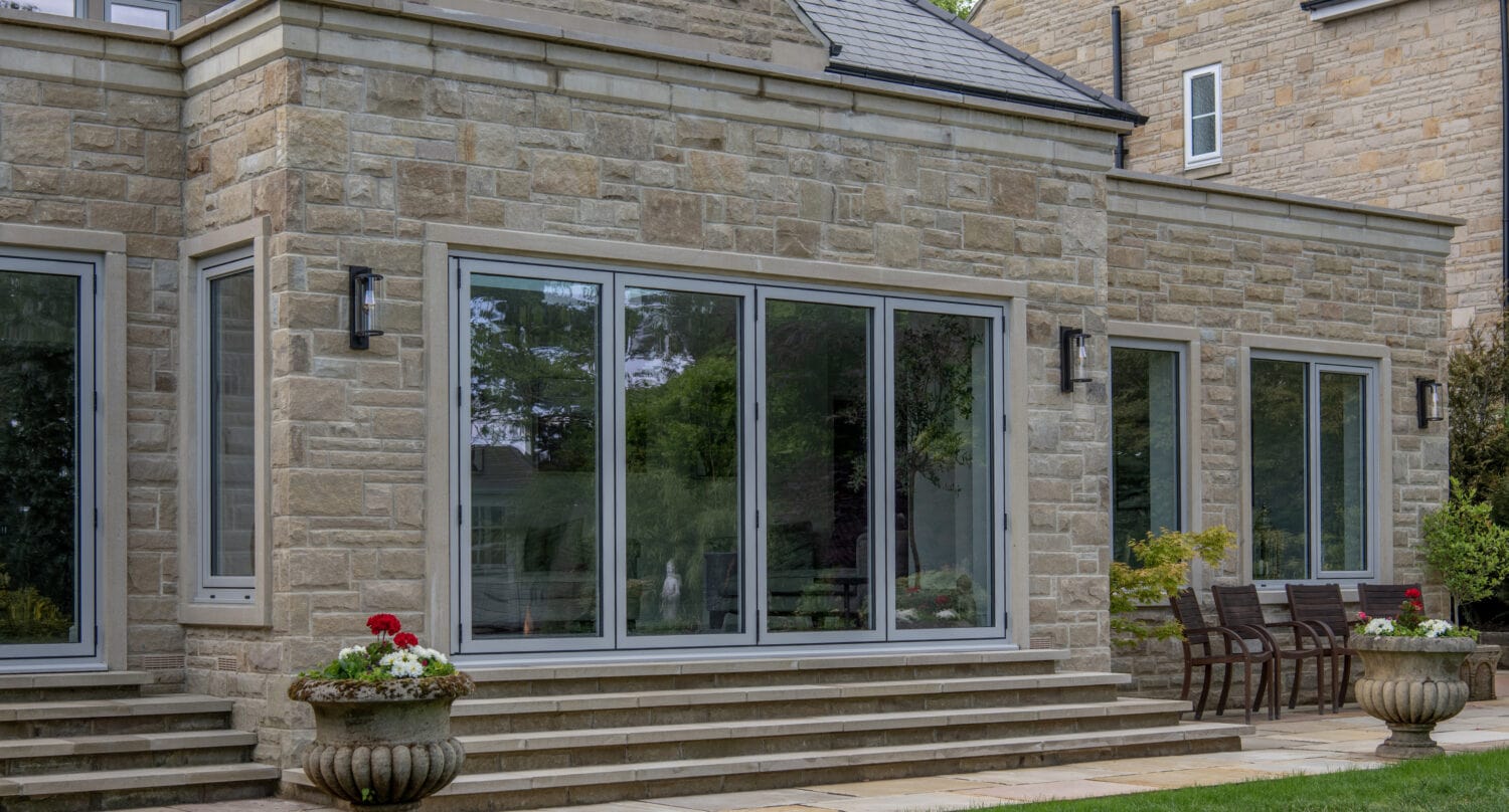 A modern house with large sliding glass doors, stone exterior, and three steps leading to a patio. Potted plants and outdoor seating are arranged beside the entrance, complemented by bespoke glass solutions for a sleek finish.